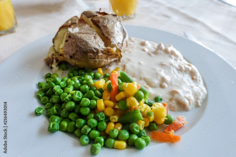 A meal of baked potato, chicken in white sauce and mixed vegetables on a white plate. A meal of baked potato, chicken in white sauce and mixed vegetables on a white plate.