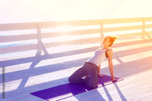 Young woman practicing yoga outdoors at white wooden seafront. Sitting in easy pose and bending back for stretching.