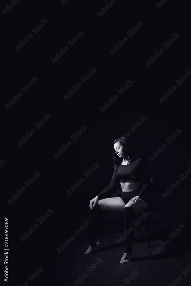 Sporty girl in black shorts and top sitting on a chair on a black isolated background black and white image. Female portrait in a low key.