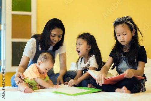 Babysitting playing with three foolish playful kids carrying on the bed; widow young woman and child are enjoying funny time indoor in modern yellow bed room.