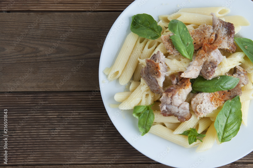 Cooked penne pasta isolated in white round plate, with basil leaves and pork meat, over wooden table, decorated with basil leaves. Top view. Cooked penne pasta isolated in white round plate, with basil leaves and pork meat, over wooden table, decorated with basil leaves. Top view.