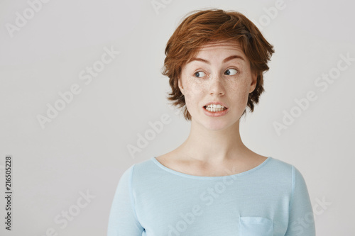 Portrait of confused attractive redhead female feeling awkward, lifting eyebrows and looking aside with weird smile after making mistake and being guilty, saying oops over gray background