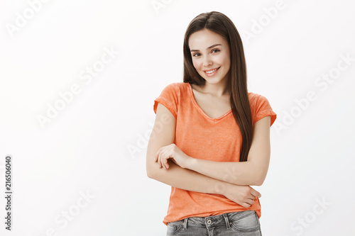 Timid cute girl asks for help. Portrait of insecure good-looking female in orange t-shirt, holding hands together on chest, smiling broadly and gazing at camera with friendly flirty expression