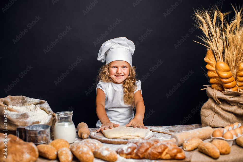 Charming little girl with curly hair in white apron and hat standing at table kneading bread dough and looking at camera. KId in good mood, having fun. Charming little girl with curly hair in white apron and hat standing at table kneading bread dough and looking at camera. KId in good mood, having fun.