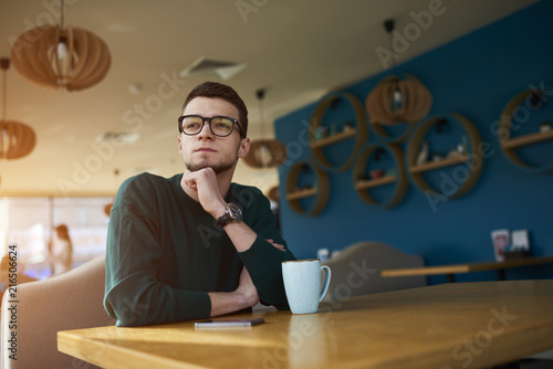 Talented young student hipster guy dressed in casual pullover looking out of window and working over tasks thinking and concentrated on developing own startup web design company photo