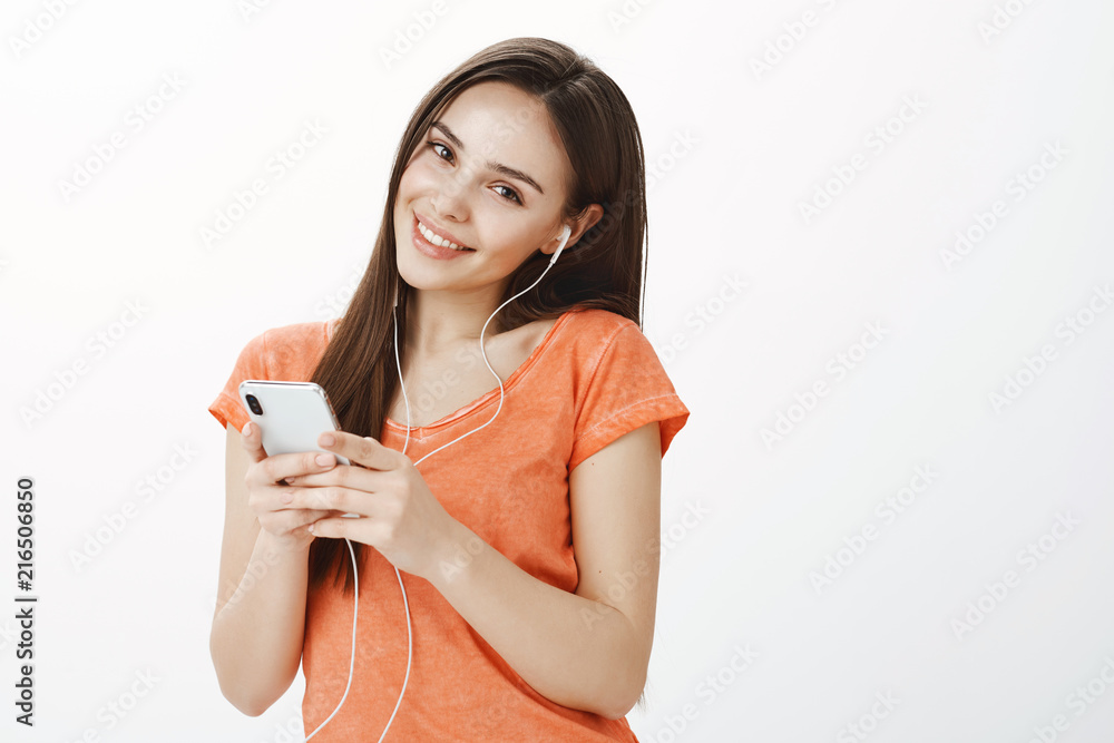 Studio shot of happy attractive caucasian female with long beautiful hair, tilting while feeling satisfaction with great bits in earphones, holding smartphone and listening music over gray background Studio shot of happy attractive caucasian female with long beautiful hair, tilting while feeling satisfaction with great bits in earphones, holding smartphone and listening music over gray background
