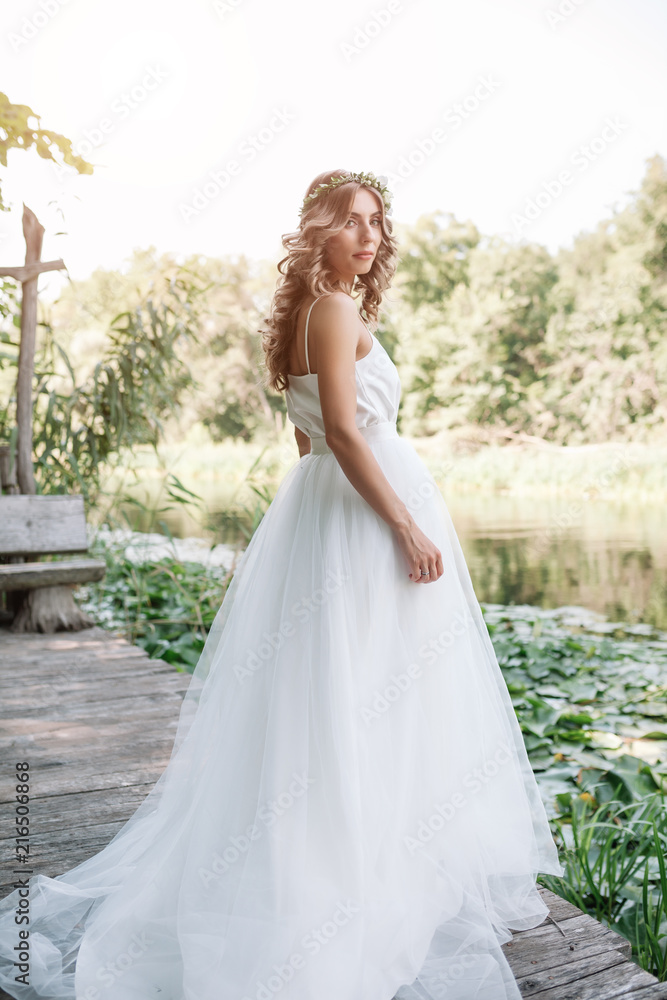 A cute curly woman in a white wedding dress with a wedding bouquet and wreath in her hair standing back to the camera in nature. Concept escaped bride. Forward to a happy bright future Runaway