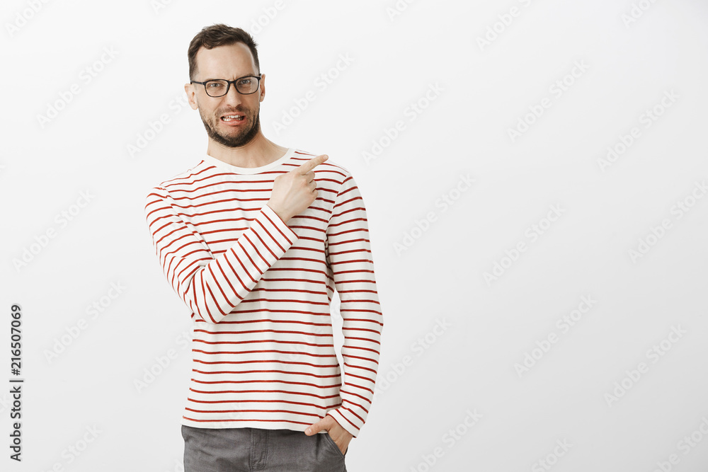 Indoor shot of disgusted displeased funny male model with bristle in black eyewear, sticking out tongue and making dislike grimace while pointing at upper right corner, expressing antipathy to item Indoor shot of disgusted displeased funny male model with bristle in black eyewear, sticking out tongue and making dislike grimace while pointing at upper right corner, expressing antipathy to item
