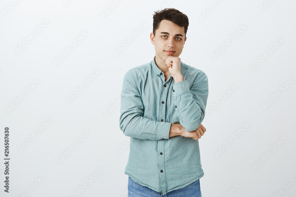 Friendly attractive european male student in turquois shirt, holding hand on chin and smiling with pleased expression, having great idea in mind, thinking while gazing at camera over gray wall Friendly attractive european male student in turquois shirt, holding hand on chin and smiling with pleased expression, having great idea in mind, thinking while gazing at camera over gray wall