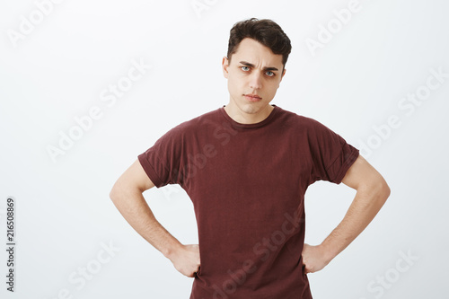 Studio shot of disappointed man boyfriend in red t-shirt, holding hands on hips, frowning and tilting head while blaming girlfriend for coming late night, being upset and scolding her over gray wall