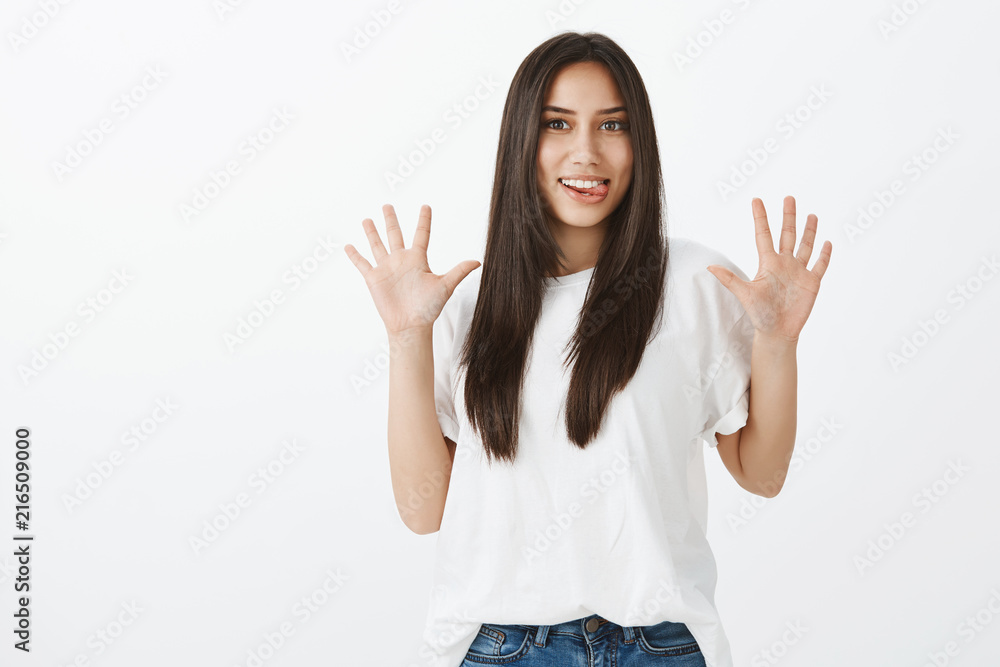 Jazz hands makes day brighter. Childish playful funny girl with long dark hair in casual outfit, raising palms and sticking out tongue, making faces while playing with sister over gray wall Jazz hands makes day brighter. Childish playful funny girl with long dark hair in casual outfit, raising palms and sticking out tongue, making faces while playing with sister over gray wall