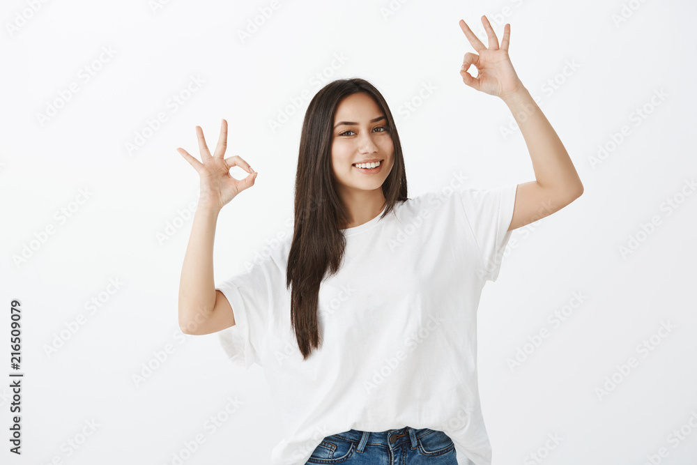 Say okay. Portrait of friendly-looking happy woman in white t-shirt and jeans, raising hands with great or acception gesture, giving approval and smiling broadly while smiling over gray wall Say okay. Portrait of friendly-looking happy woman in white t-shirt and jeans, raising hands with great or acception gesture, giving approval and smiling broadly while smiling over gray wall