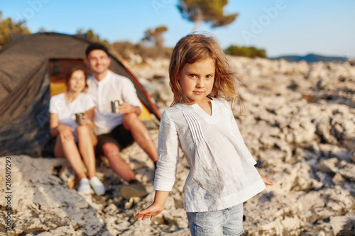 Little girl with brown eyes, standing on rocky beach and looking at camera.