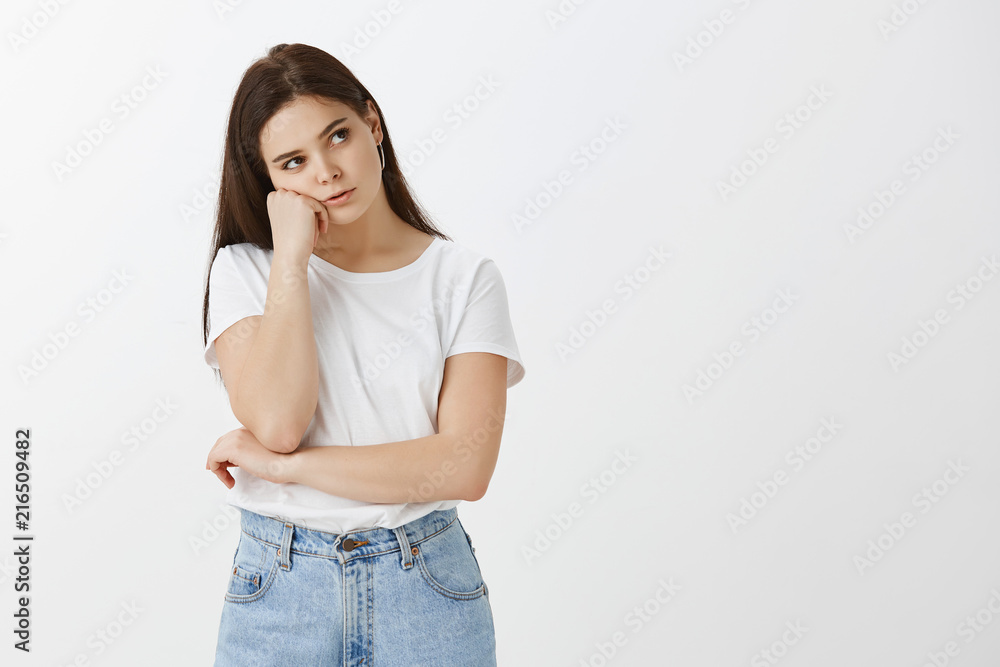 Studio shot of sad bored cute woman in t-shirt and jeans, leaning on fist and gazing up while checking time during boring lesson, standing tired and upset over gray background, wanting to have fun Studio shot of sad bored cute woman in t-shirt and jeans, leaning on fist and gazing up while checking time during boring lesson, standing tired and upset over gray background, wanting to have fun