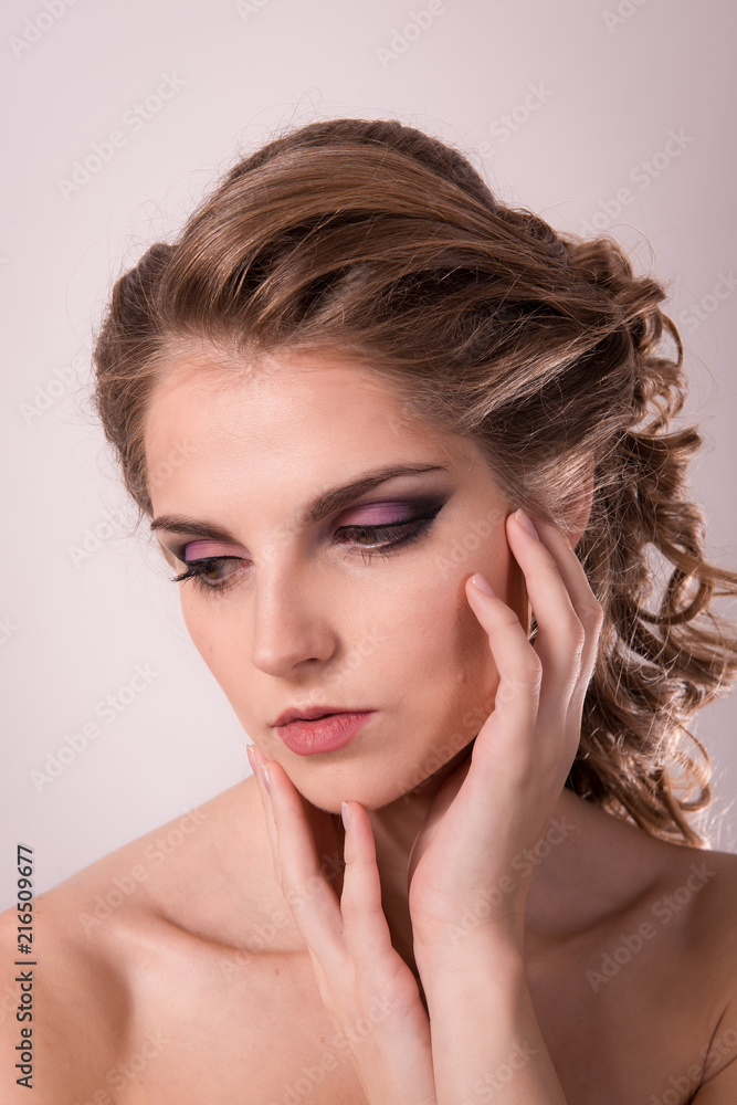 Brown-haired woman with a professional hairdress and make-up is holding hands near the face on a light background close-up. Brown-haired woman with a professional hairdress and make-up is holding hands near the face on a light background close-up.