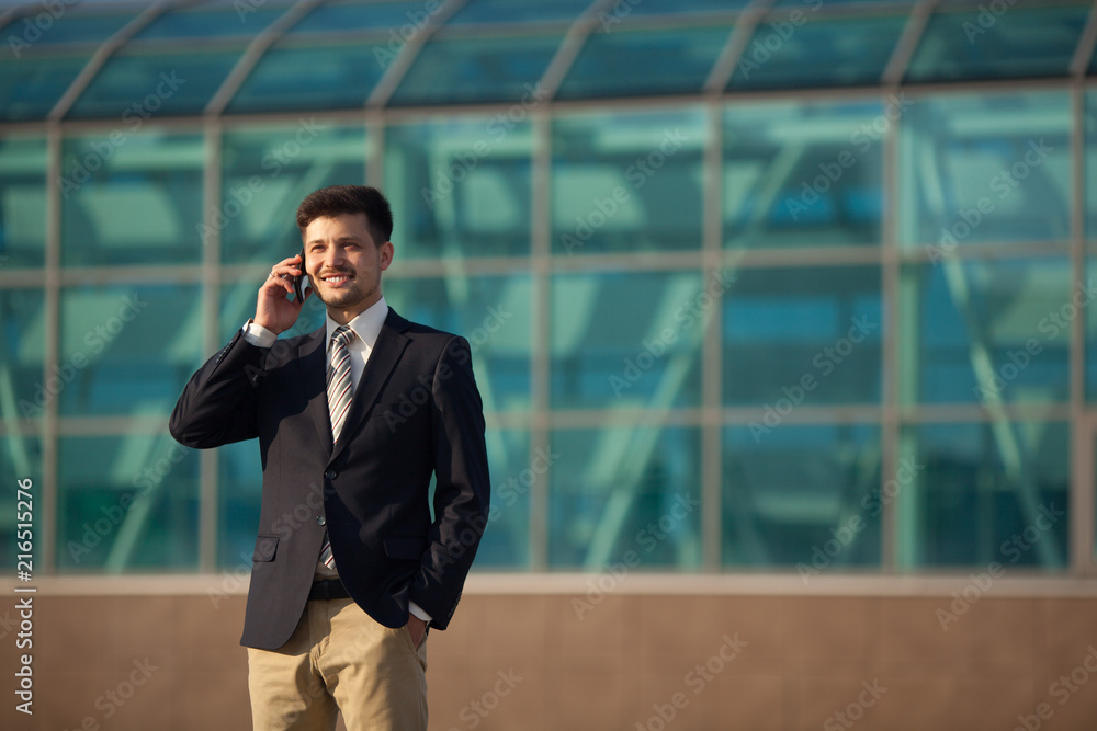 handsome young man in a suit standing near a glass building talking on the phone in the summer handsome young man in a suit standing near a glass building talking on the phone in the summer
