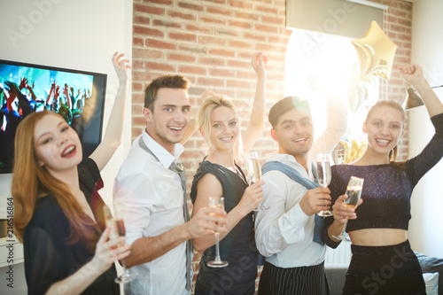 Row of ecstatic young people in smart attire raising flutes of champagne while having fun at home party