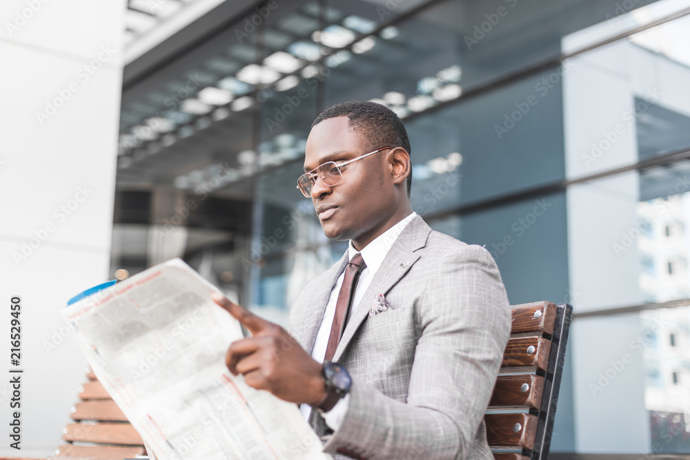 black man businessman in a business suit and glasses sits on a bench and reads a newspaper against the backdrop of a modern city black man businessman in a business suit and glasses sits on a bench and reads a newspaper against the backdrop of a modern city