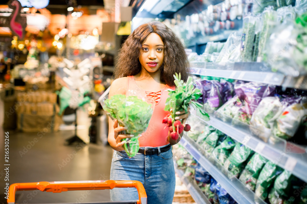 Pretty Afro young woman in jeans shopping organic veggies and fruits. Woman with trolley holding lettuce and radish in her arms