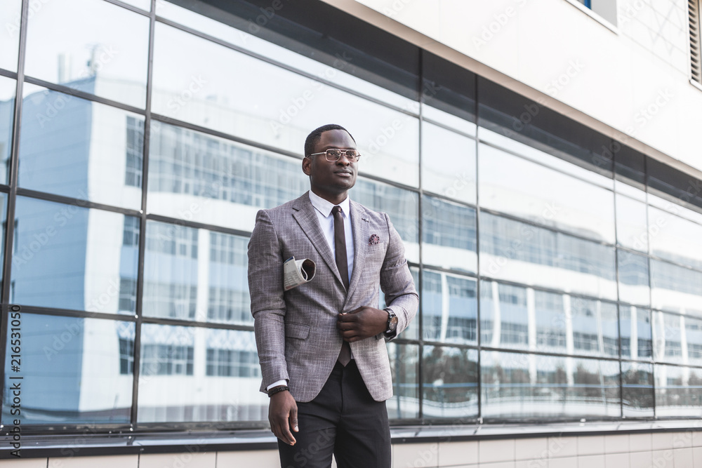 black man businessman in a business suit, expensive watch and glasses with a newspaper goes against the backdrop of a modern city to work black man businessman in a business suit, expensive watch and glasses with a newspaper goes against the backdrop of a modern city to work