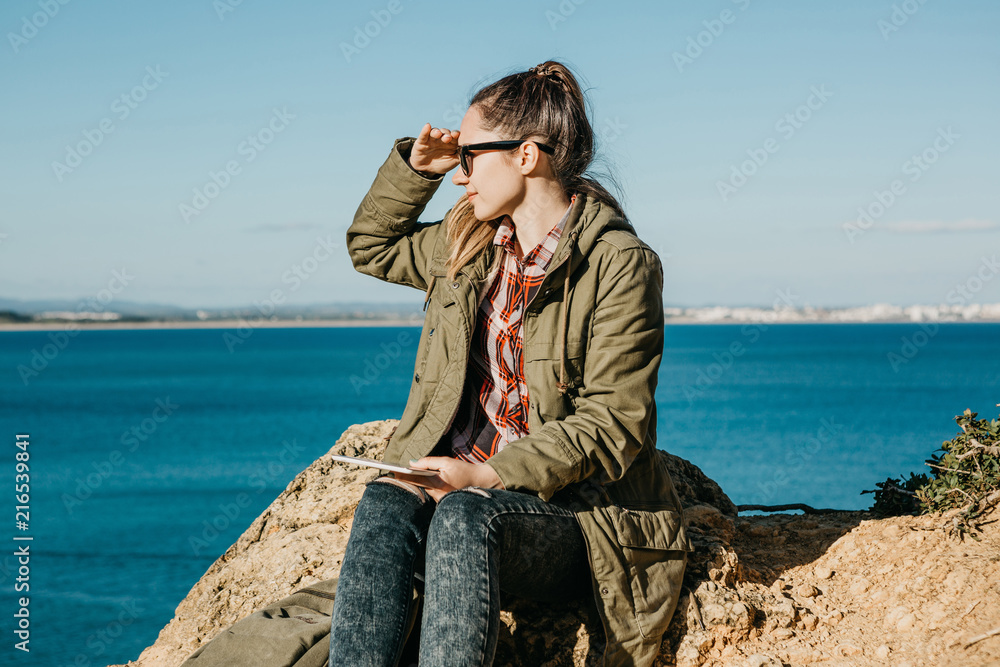 A young beautiful girl or tourist uses a tablet and looks into the distance. Ocean or sea in the background.