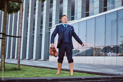 Obraz na plátne Confident businessman dressed in elegant suit holds his shoes enjoying while standing barefoot on a green lawn against cityscape background