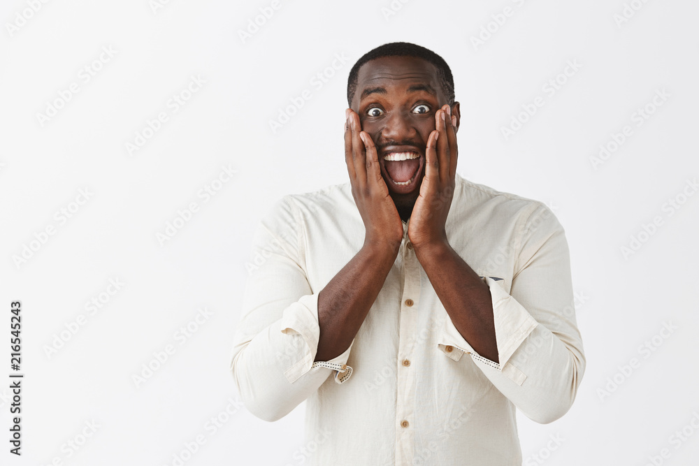 Man hearing out great news, being joyful and amazed, happy for friend. Portrait of impressed and surprised upbeat African American guy in white shirt smiling broadly and holding palms on cheeks Man hearing out great news, being joyful and amazed, happy for friend. Portrait of impressed and surprised upbeat African American guy in white shirt smiling broadly and holding palms on cheeks
