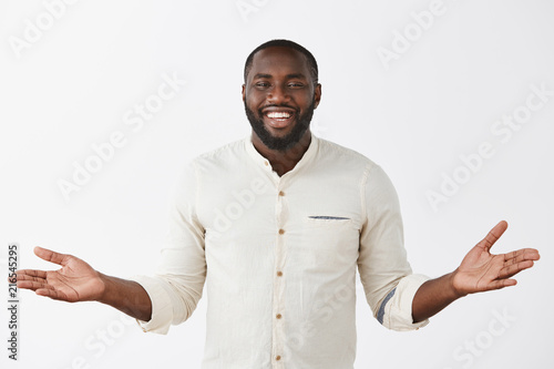 Indoor shot of charming carefree and happy cute dark-skinned bearded man in white shirt spreading palms aside and smiling broadly, inviting friend to come in, being hospitable and polite