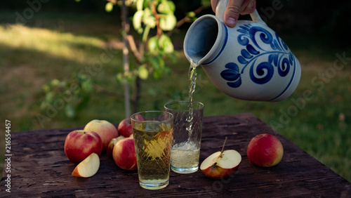 a man's hand pours Traditional apple wine in a refilled glass in the city of Frankfurt. A jug of wine on an old wooden table in the garden, around it apples photo