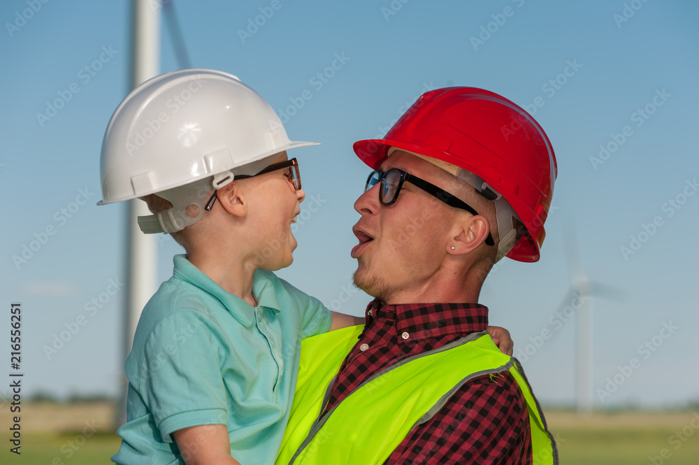 Engineer in the second generation. Portrait of a little positive boy in the hands of his father's engineer's in glasses and helmets against the background of windmill and blue sky Engineer in the second generation. Portrait of a little positive boy in the hands of his father's engineer's in glasses and helmets against the background of windmill and blue sky