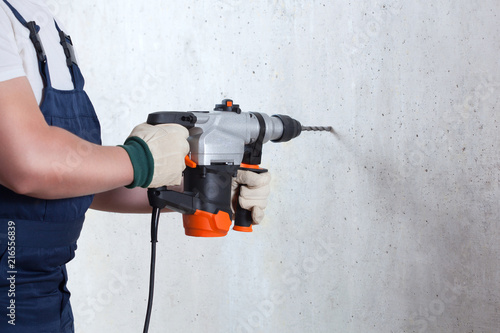 The builder with hammer drill perforator equipment making hole in wall at construction site.