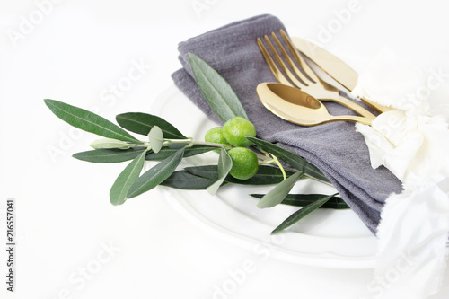 Closeup of festive table summer setting with golden cutlery, olive branch, grey linen napkin, porcelain dinner plate and silk ribbon on white table background. Mediterranean wedding, restaurant.