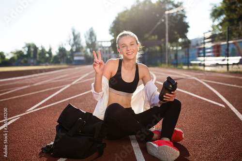 Cheerful girl in wireless earphones holding sport bottle in hand happily looking in camera while showing two fingerss gesture on running track of stadium photo