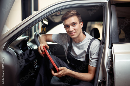 A young smiling automechanic is sitting in a car with a special device for cheching the computer system of a car