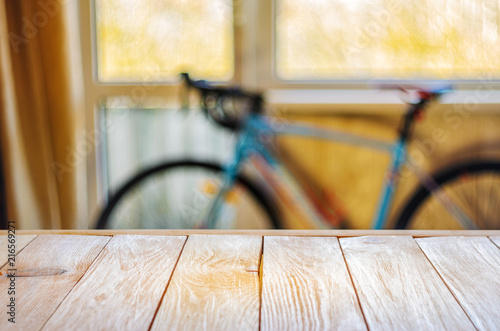 Empty Wooden Table in Front of Blurred Background of Living Room with a Blue Bicycle Near the Window. Mock Up for Display of Product. Blank for Your Layout