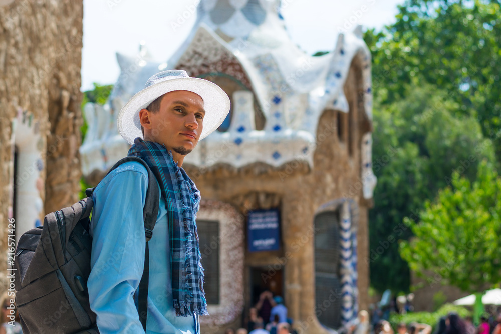 A young guy stands in a white hat and scarf with backpack near a gingerbread house in Guell Park, inside