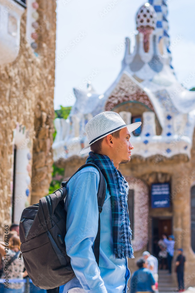 A young guy stands in a white hat and scarf with backpack near a gingerbread house in Guell Park, inside A young guy stands in a white hat and scarf with backpack near a gingerbread house in Guell Park, inside