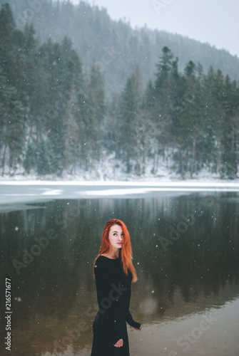 Handsome redhead girl looking up on the snowflakes falling on her hair. Splendid shady black mountain lake. Dreamy winter landscape of Carpathian mountains.