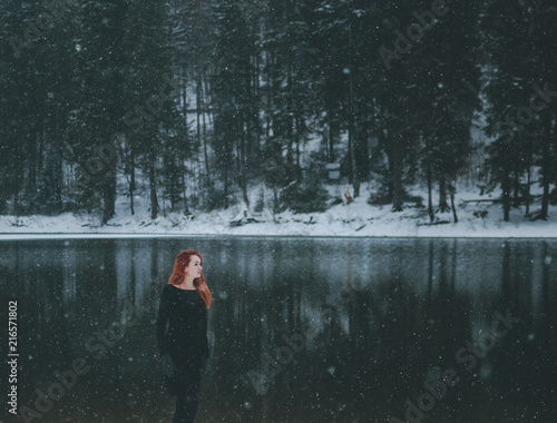 Snowflakes flying over handsome black-eyed redhead girl looking in the distance. White snowflakes flying all around. Splendid shady black mountain lake