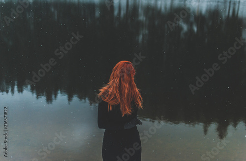 Snowflakes flying over handsome black-eyed redhead girl looking in the distance. White snowflakes flying all around. Splendid shady black mountain lake. Look from behind