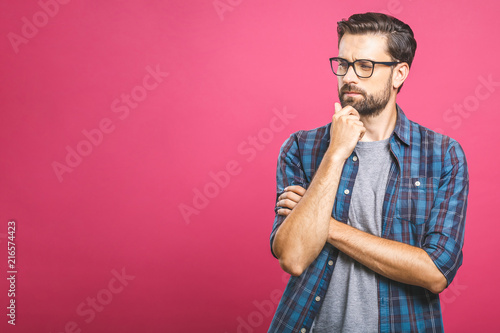 Portrait of unhappy determined european male with bristle touching chin while thinking and looking with serious and worried look at camera, standing against pink background. photo