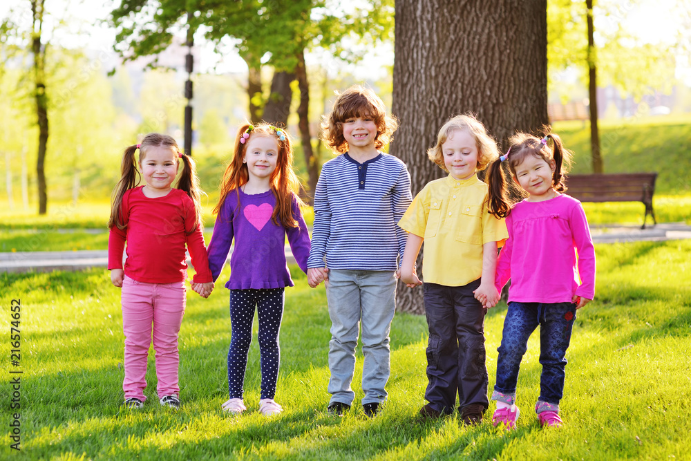 a group of small children smiling holding hands on a background of grass, a tree and a park. Children's Day, June 1, friendship, childhood. a group of small children smiling holding hands on a background of grass, a tree and a park. Children's Day, June 1, friendship, childhood.