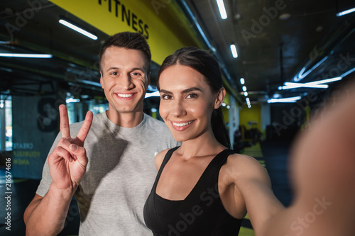 Fototapeta Naklejka Na Ścianę i Meble -  Portrait of satisfied man and woman taking their own photo after workout. Smiling girl is holding gadget for shooting while guy is grinning and expressing v gesture. Enjoying active time with partner