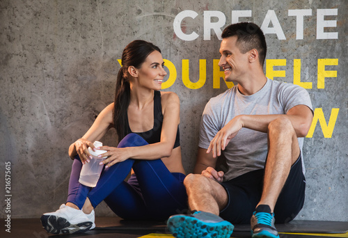 Positive guy is coGrinning couple is relaxing and sitting on floor in sport center after workout. They are having pleasure while chatting and resting. Woman is holding flask with water for quenching