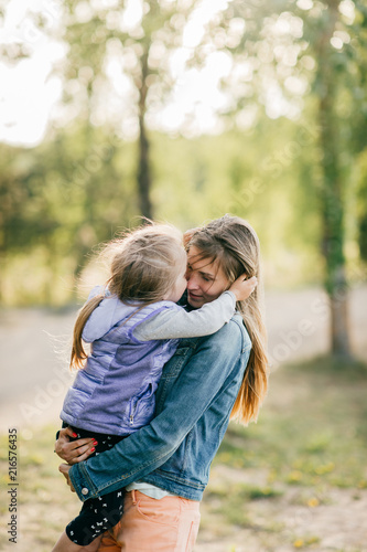 Happy smiling mother hugging her lovely little daughter outdoor. Lifestyle family. Adult cheerful female parent playing with her beautiful emotional child at nature in summer. Positive people faces.