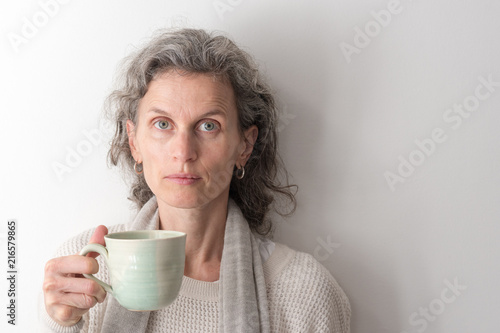 Portrait of middle aged woman with grey hair holding green cup and looking directly at camera against neutral wall background  selective focus 
