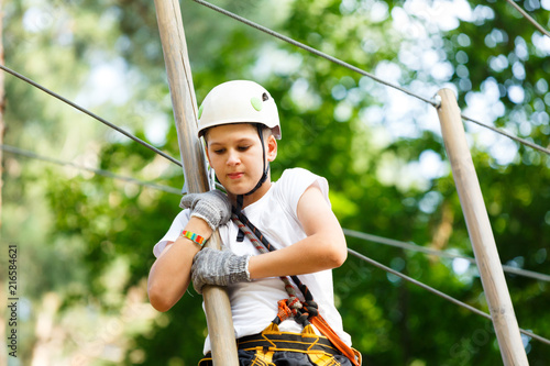Happy, cute, young boy in white t shirt and helmet having fun and playing at adventure park, holding ropes and climbing wooden stairs. active lifestyle concept