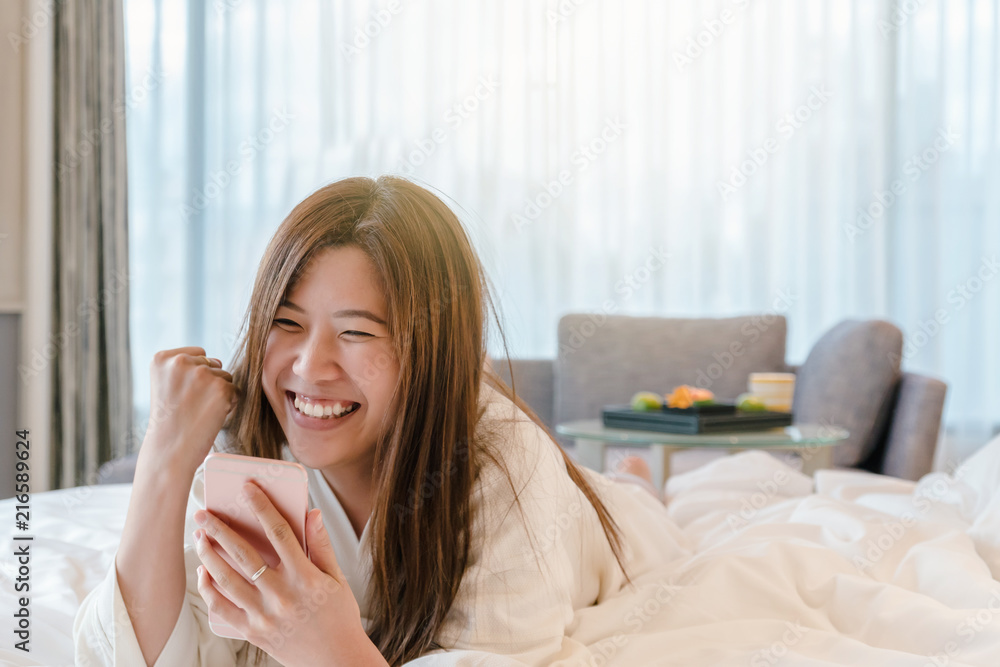 Asian woman in bathrope suit using the smart mobile phone with success action on the bed when wake up in luxury hotel, lifestyle and leisure concept Asian woman in bathrope suit using the smart mobile phone with success action on the bed when wake up in luxury hotel, lifestyle and leisure concept