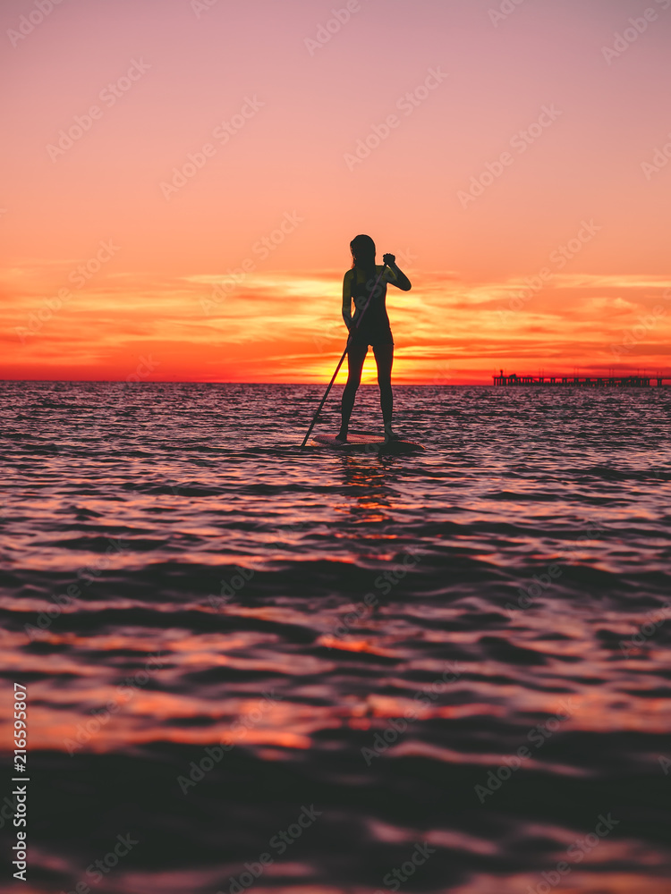 Sporty woman stand up paddle boarding  on a flat warm quiet sea with bright sunset