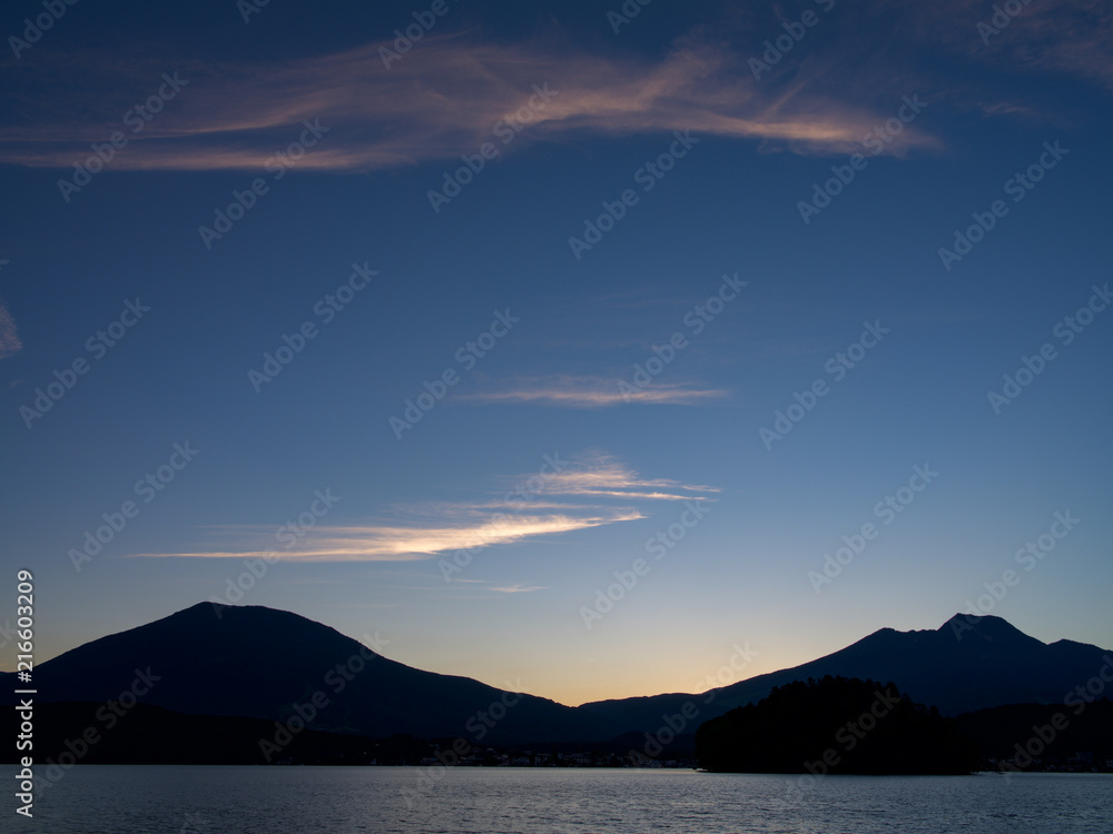 夕焼けの湖畔にて、山の間に太陽が沈み青い空に雲がなびく